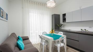 A modern kitchen with white cabinetry, a dining table with chairs, and a grey sofa with teal cushions.