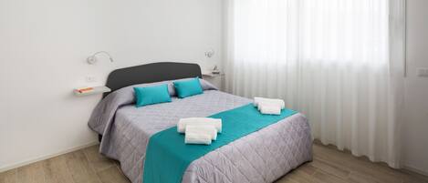 A neatly made bed with a teal blanket and pillows, a bedside table with a lamp, and a window with sheer curtains.