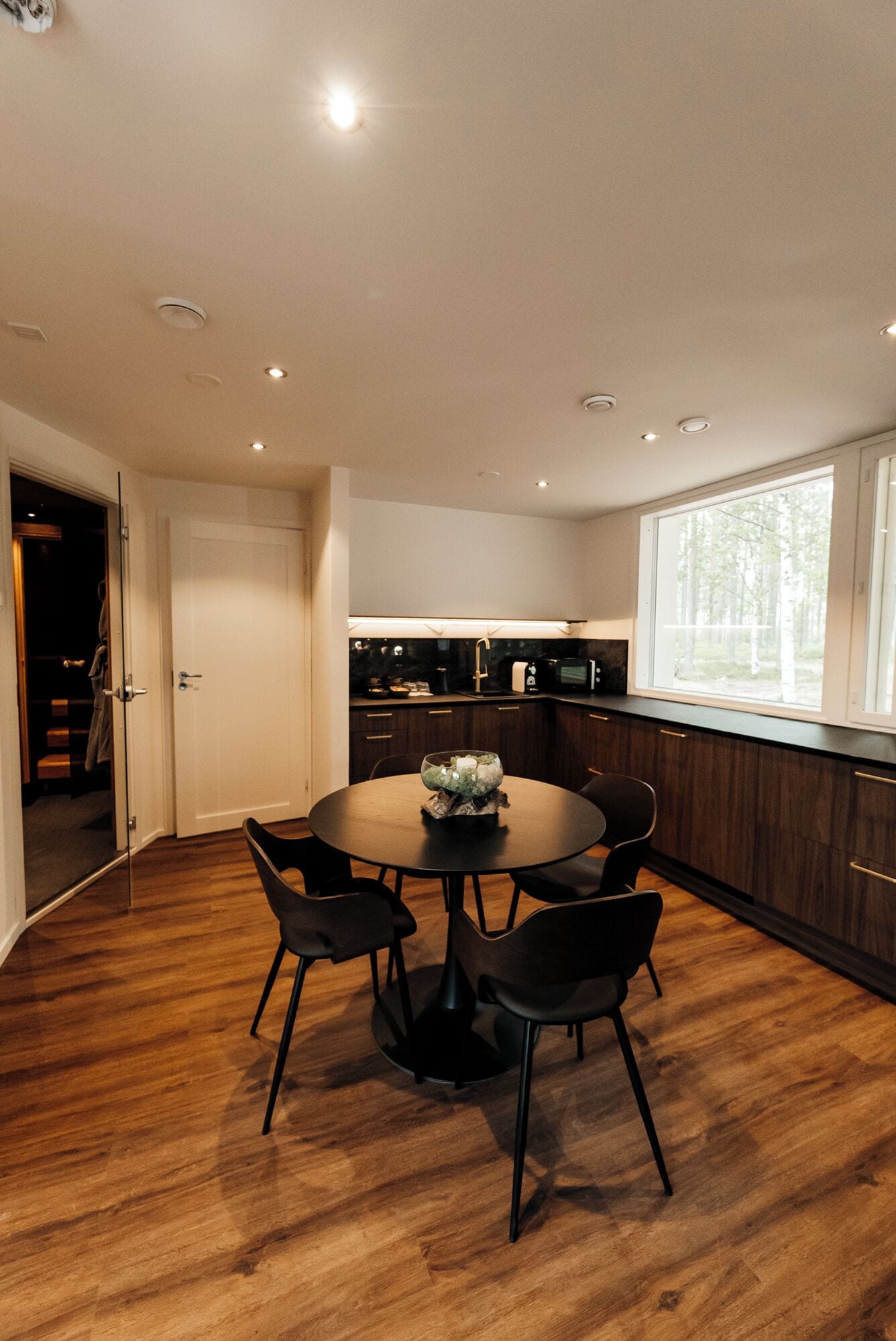 A modern kitchen with dark wood cabinets, a round dining table with four chairs, and a large window providing natural light.