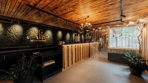 A spa reception area with a wooden ceiling, a stone wall with a logo, a chandelier, and a window with a view of trees. - Aito  Igloo & Spa Resort (Rovaniemi)