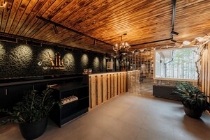 Zona de recepción de un spa con techo de madera, pared de piedra con un logotipo, una araña y una ventana con vistas a árboles.