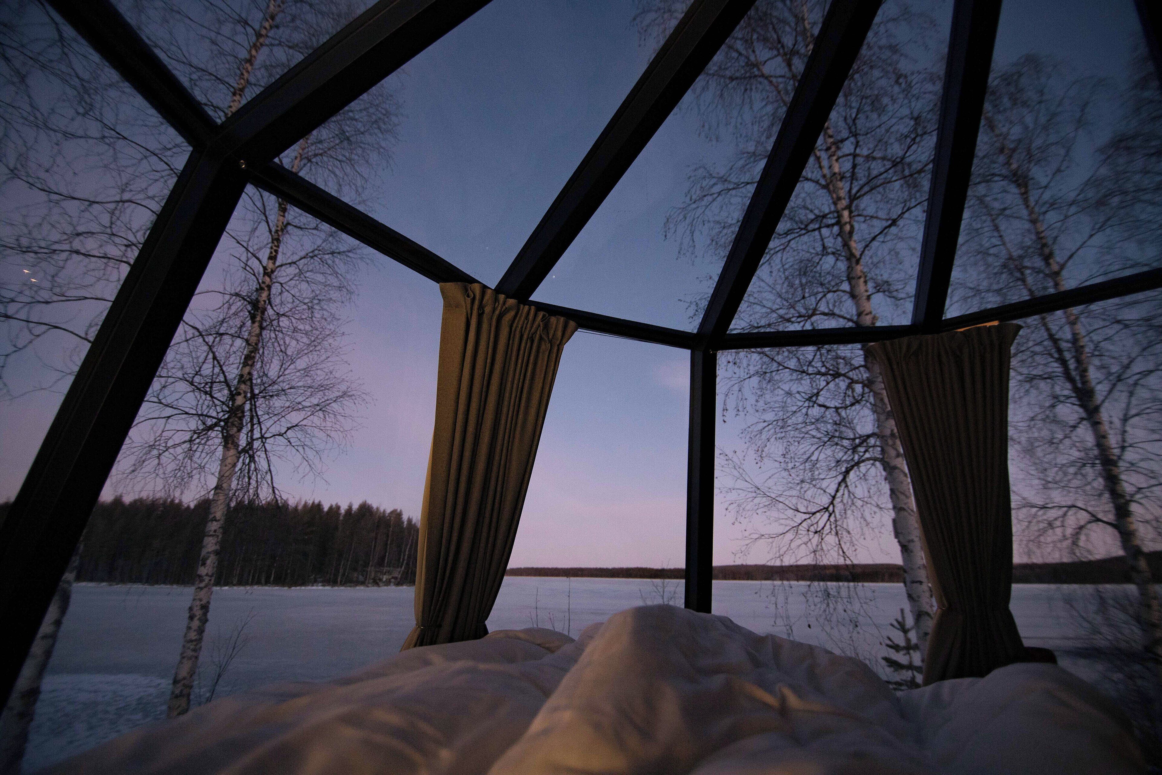 Huoneessa on suuri ikkuna, josta avautuu näkymä jäiselle järvelle ja puille, ja etualalla on sänky.