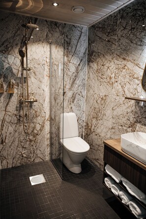 A modern bathroom with a marble wall, a glass-enclosed shower, a white toilet, and a wooden vanity with a marble sink.