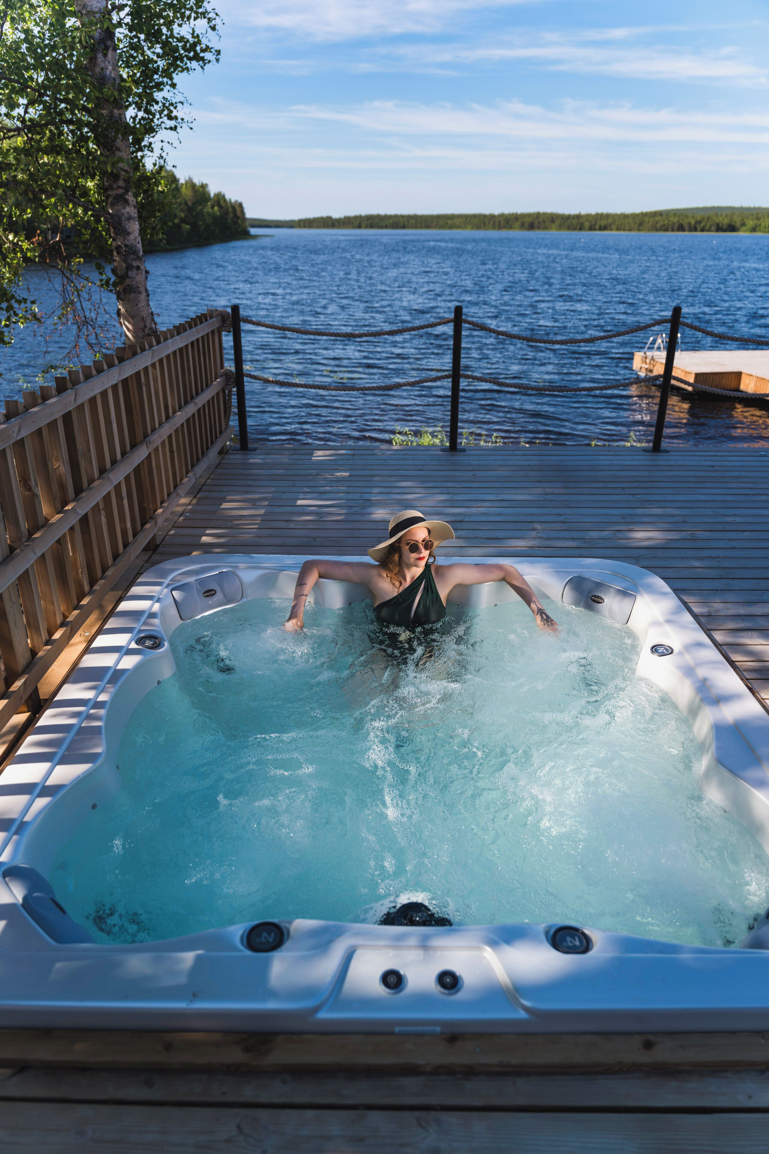 A person relaxing in a hot tub on a wooden deck by a lake.