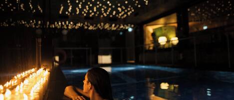 Une femme se trouve dans un espace intérieur faiblement éclairé, près d’une piscine, avec des bougies disposées le long du bord.