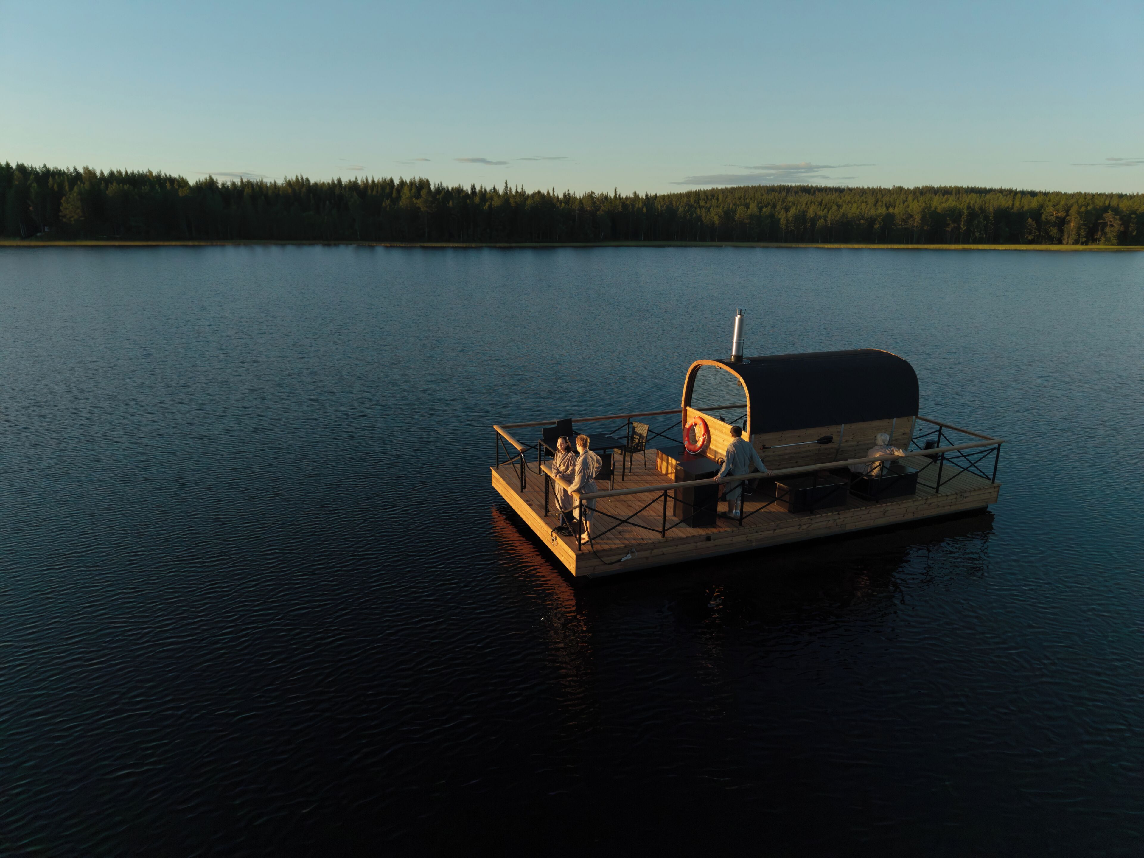 A stationary boat with a covered deck, seating, and a chimney, floating on calm water with a forested shoreline in the background.