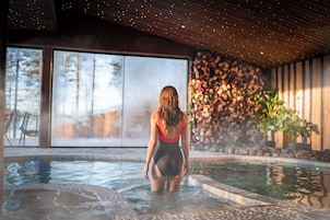 Una mujer en traje de baño de pie en un jacuzzi con un ventanal grande y un muro floral de fondo.