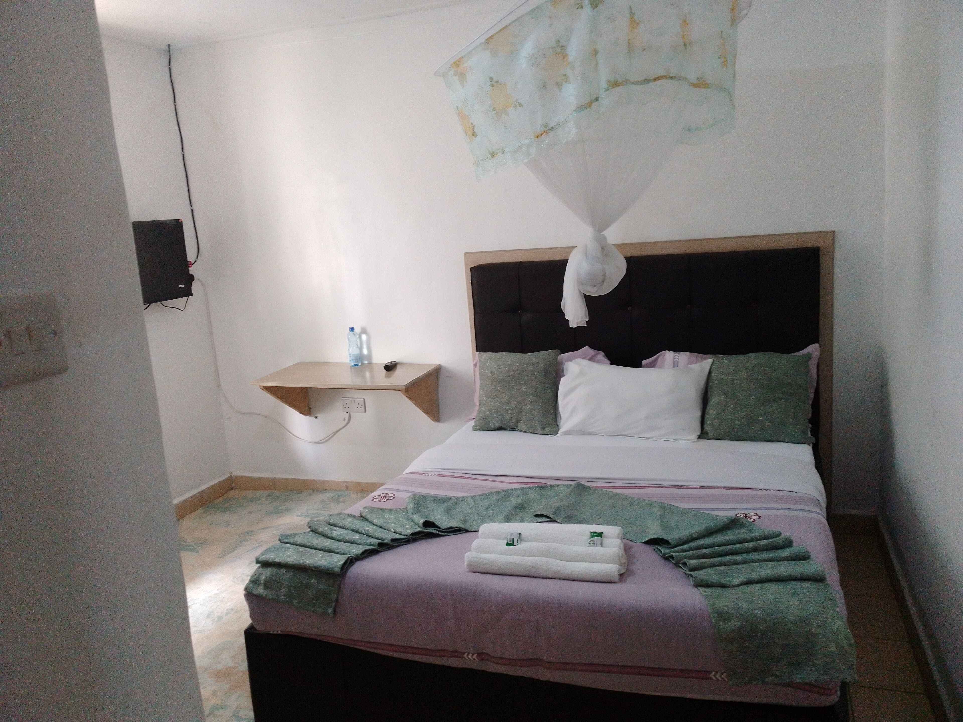A bedroom with a bed, a bedside table, a hanging mosquito net, and a wall-mounted TV.