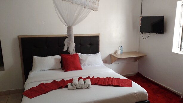 A neatly made bed with white and red bedding, a black headboard, and a canopy overhead. A flat-screen TV is mounted on the wall to the right. - WHITE HOUSE LOUNGE & GUEST HOUSE (Eldoret)
