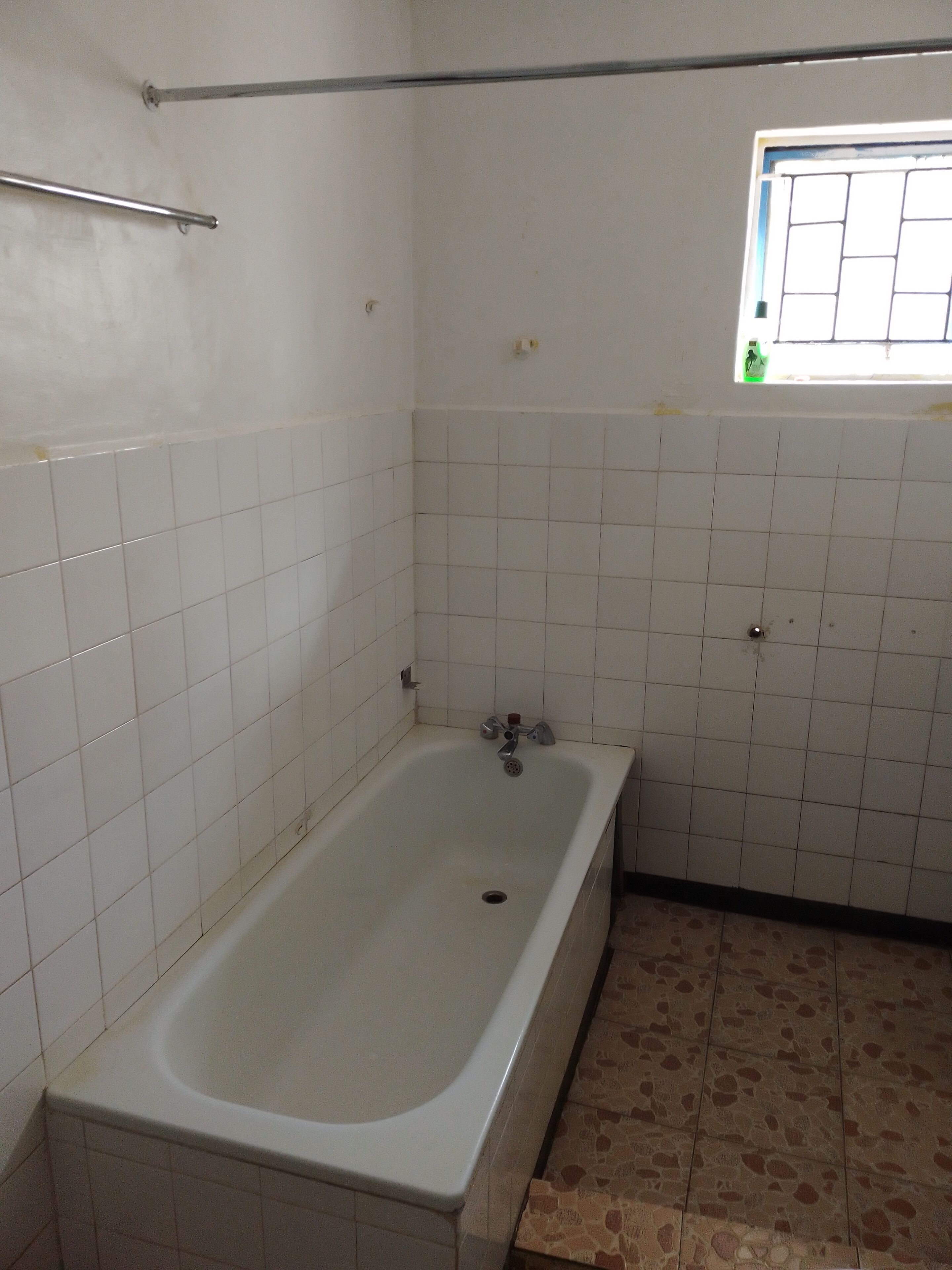 A bathroom with a bathtub, white tiled walls, a window with bars, and a tiled floor.