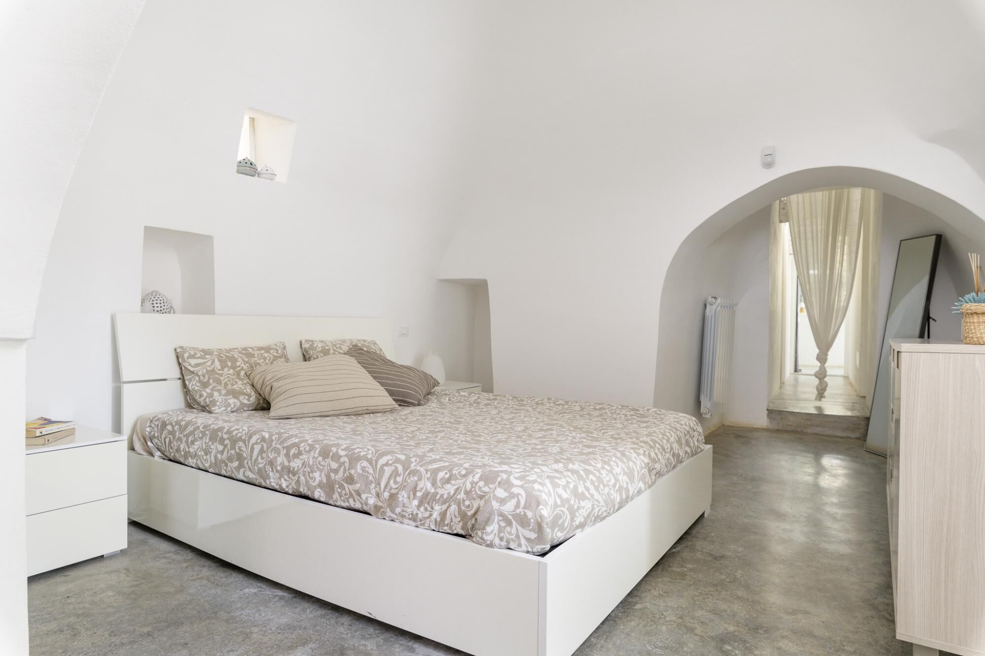 A bedroom with a bed, a nightstand, a dresser, and an arched doorway leading to another room.