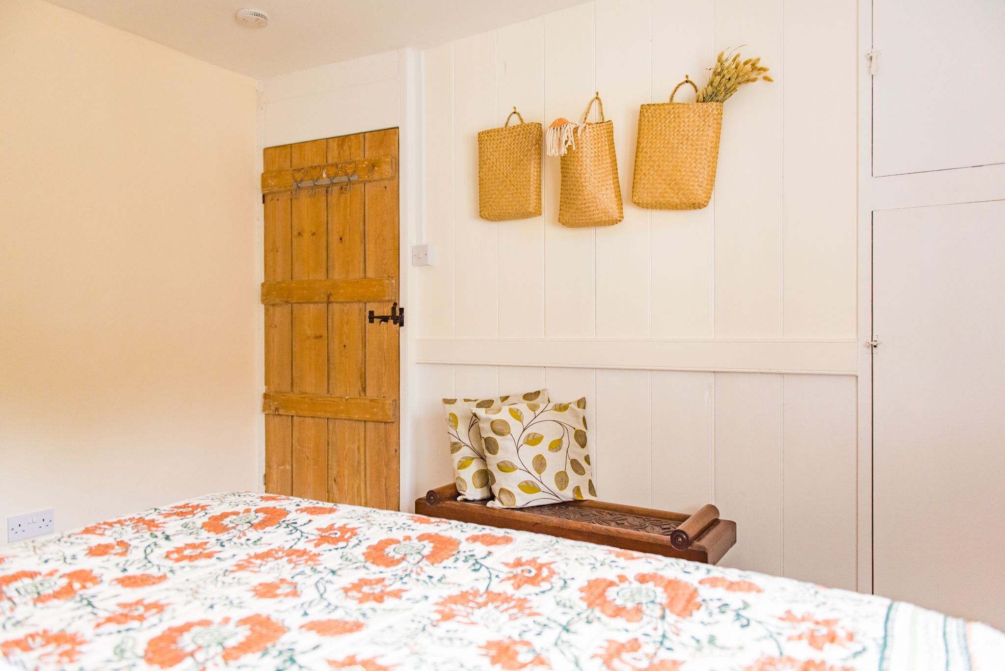 Un dormitorio con puerta de madera, una cama con colcha floral y tres cestas tejidas colgadas en la pared.