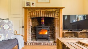 A cozy room with a brick fireplace, a television, and a wooden table. - Hearts Delight Cottage by Bloom Stays -  a holiday let that sleeps 4 guests  in 2 bedrooms (Canterbury)