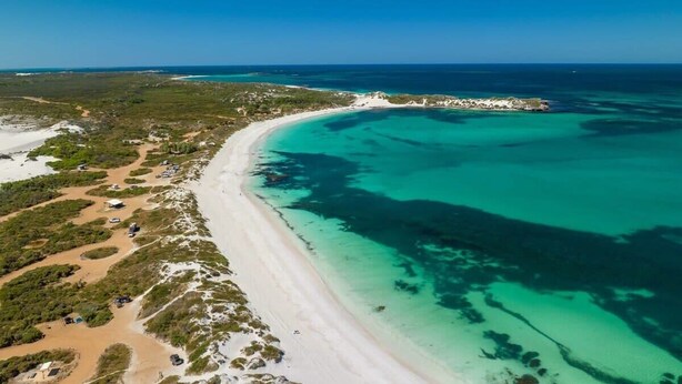Vue aérienne d’une zone côtière avec une plage de sable, des eaux turquoise et limpides, et une végétation clairsemée.