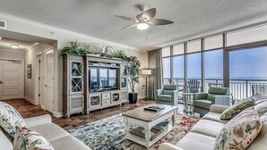 A spacious living room with a large TV, comfortable seating, a ceiling fan, and a view of the ocean.