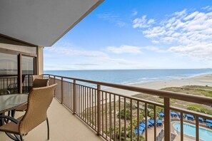 A balcony with a view of the ocean, a pool area with blue umbrellas, and a dining table with chairs.