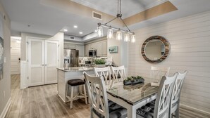 Una cocina moderna con armarios blancos, una isla central y una zona de comedor con una mesa de vidrio y sillas blancas.