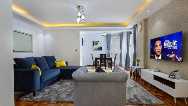 A modern living room with a sectional sofa, a grey armchair, a dining area with a table and chairs, and a TV mounted on the wall. - Nairobi's Oasis: Pool, Gym, & Modern Comforts (Nairobi)