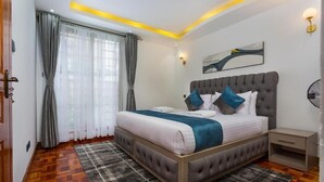 A bedroom with a large bed, a bedside table, a wall-mounted light, and a window with curtains. - Nairobi's Oasis: Pool, Gym, & Modern Comforts (Nairobi)