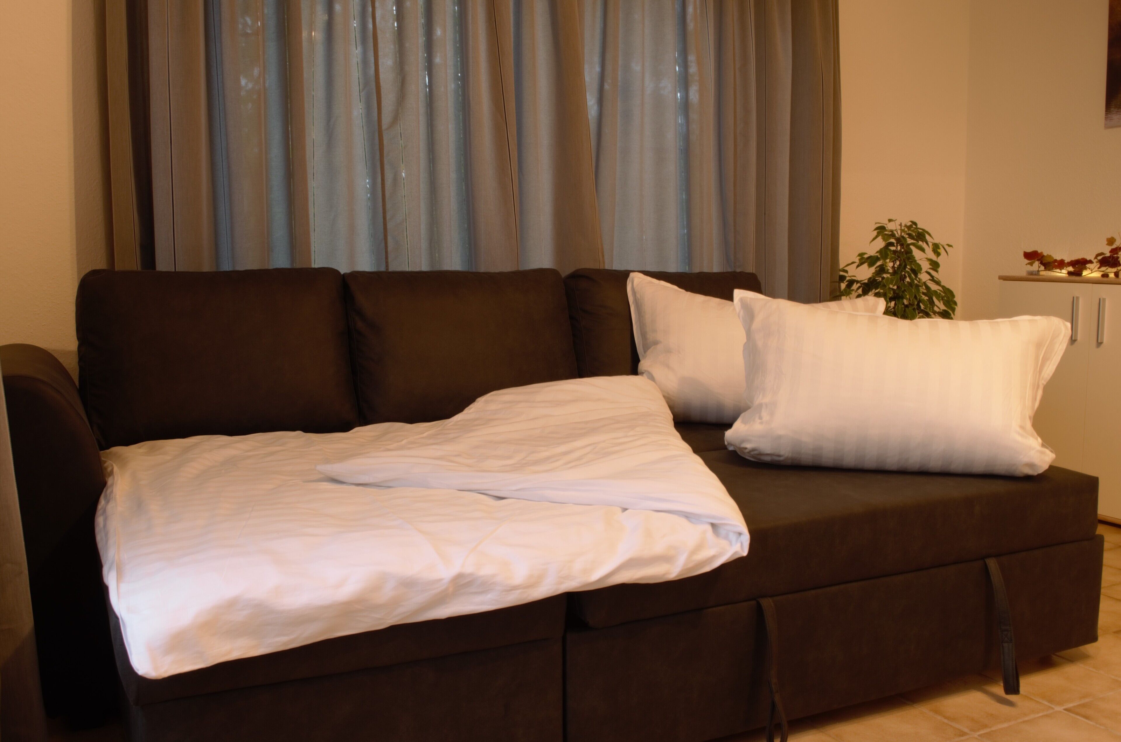 Un canapé brun foncé, agrémenté de coussins blancs et recouvert d’une couverture blanche, est placé devant une fenêtre ornée de rideaux bleus.