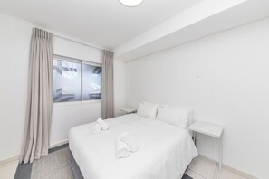 A hotel room with a large bed, a nightstand, a window with curtains, and a view of palm trees.