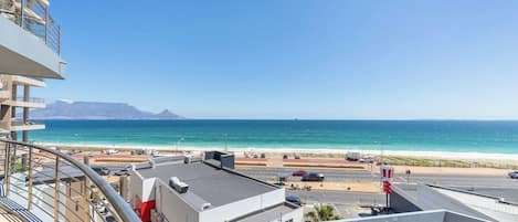 A balcony with a view of the beach and ocean, a parking area, and a clear sky.