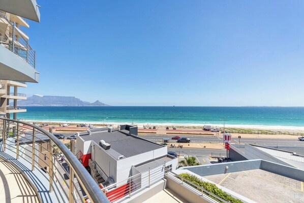 A balcony with a view of the beach and ocean, a parking area, and a clear sky.