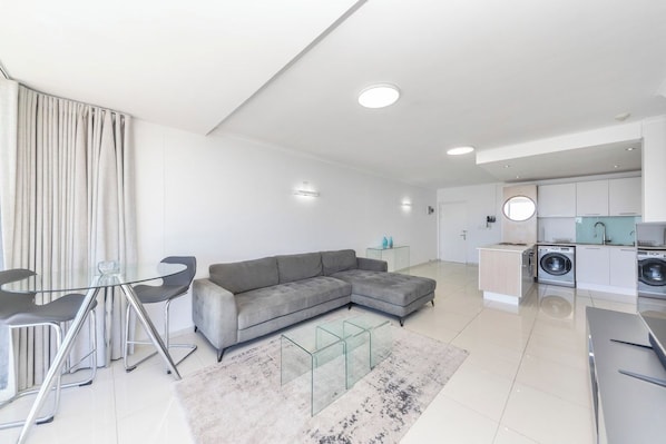 A modern living room with a grey sectional sofa, a glass coffee table, and a dining area with a glass table and metal chairs.