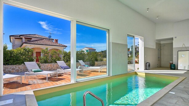 Une piscine intérieure moderne, avec une eau d’un bleu limpide, de grandes portes-fenêtres coulissantes et une vue sur une maison traditionnelle.