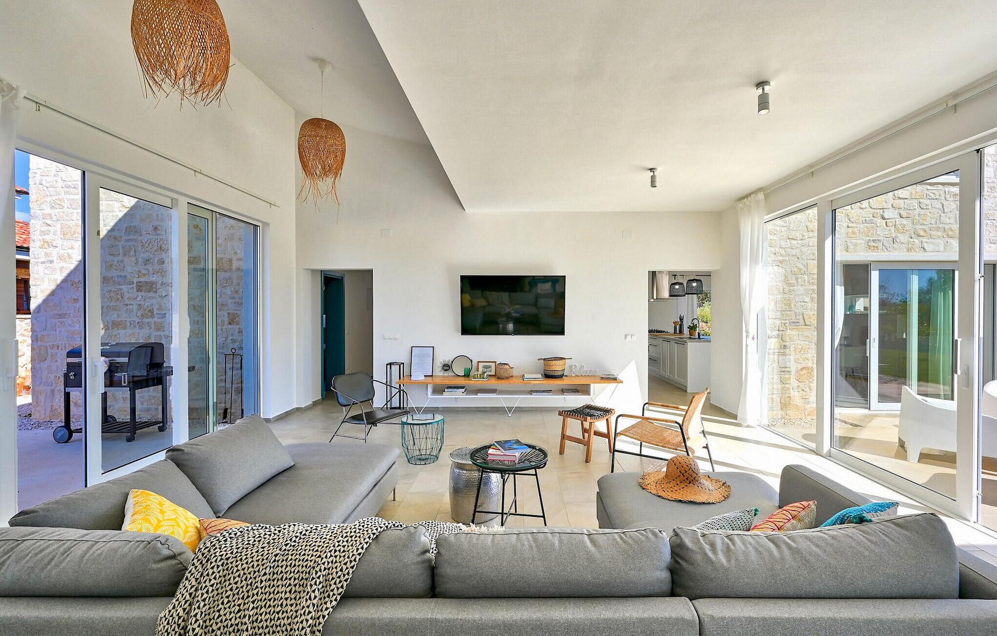 Ein großzügiger Wohnbereich mit grauem Sofa, einem Couchtisch und einem an der Wand montierten Fernseher. Es gibt Hängeleuchten aus Weidengeflecht, eine durch eine offene Tür sichtbare Küche und eine Steinwand.