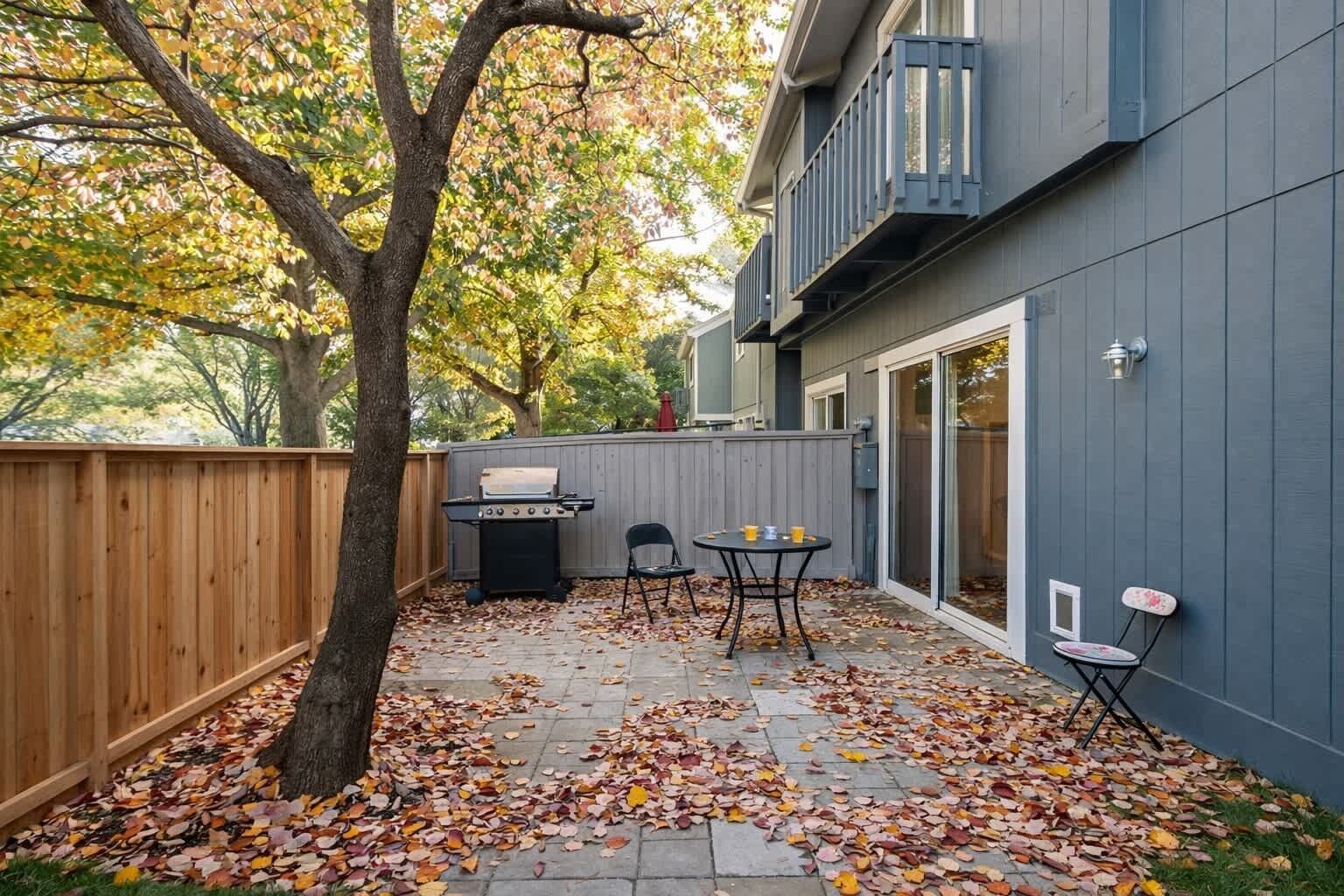 Un jardin avec un barbecue, une table et des chaises, le tout entouré d’une clôture en bois et d’un grand arbre.