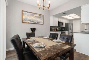 A dining area with a marble table, black leather chairs, and a chandelier. There is a kitchen in the background with modern appliances.