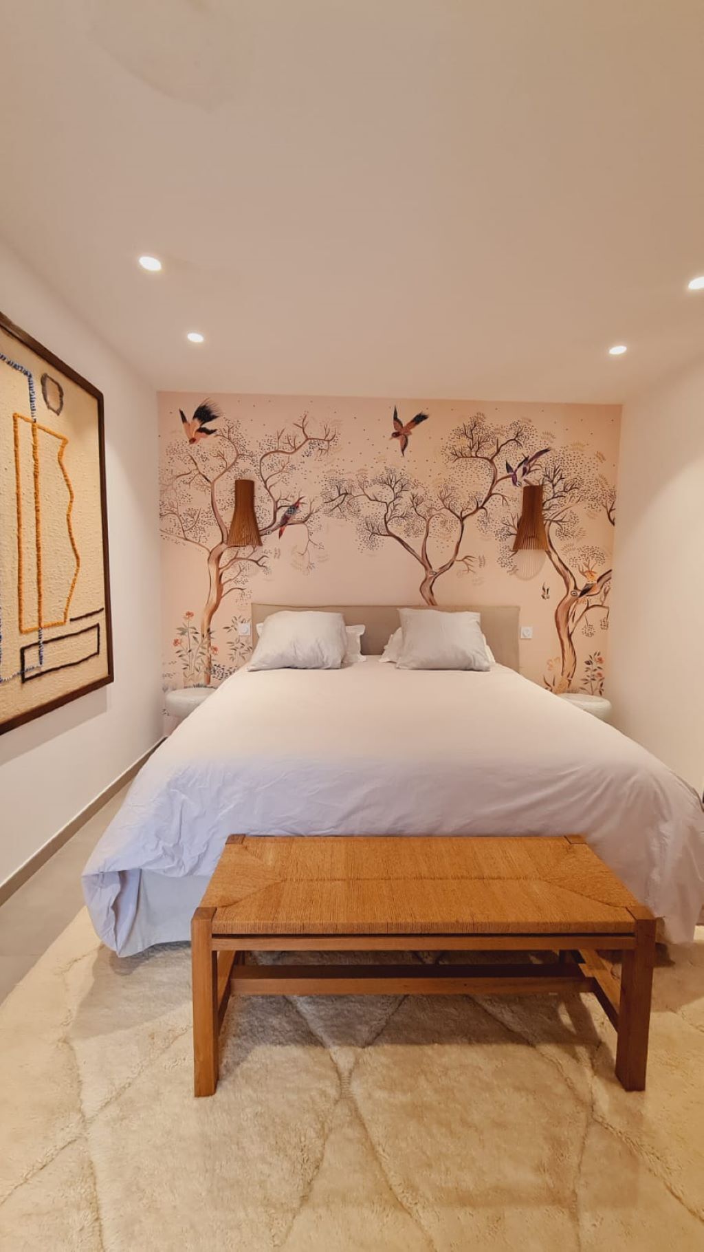 Quarto com uma cama grande, um banco de madeira e um mural na parede com árvores e pássaros.
