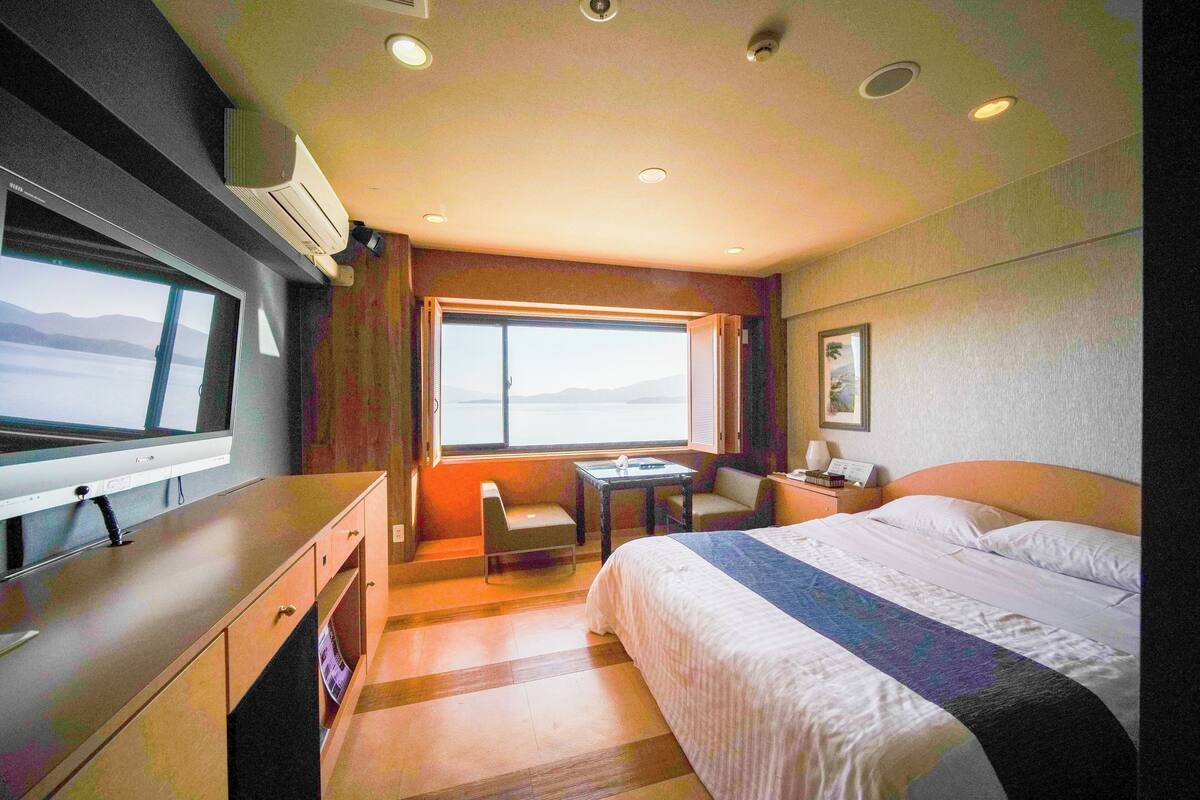 Quarto de hotel com cama, escrivaninha, cadeira, janela com vista e televisão.
