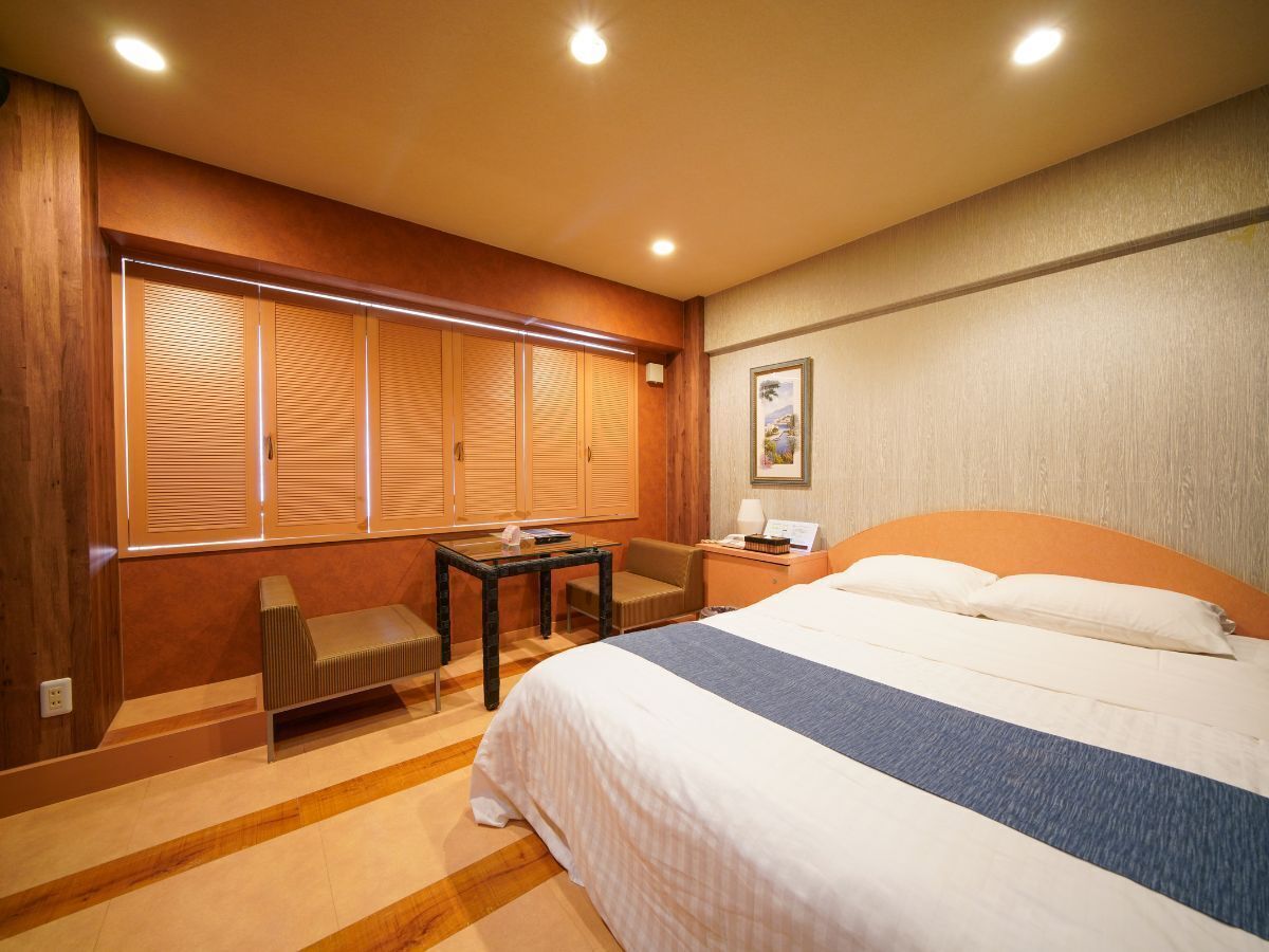 Une chambre d’hôtel avec un lit, un bureau, une chaise et une fenêtre avec des stores.