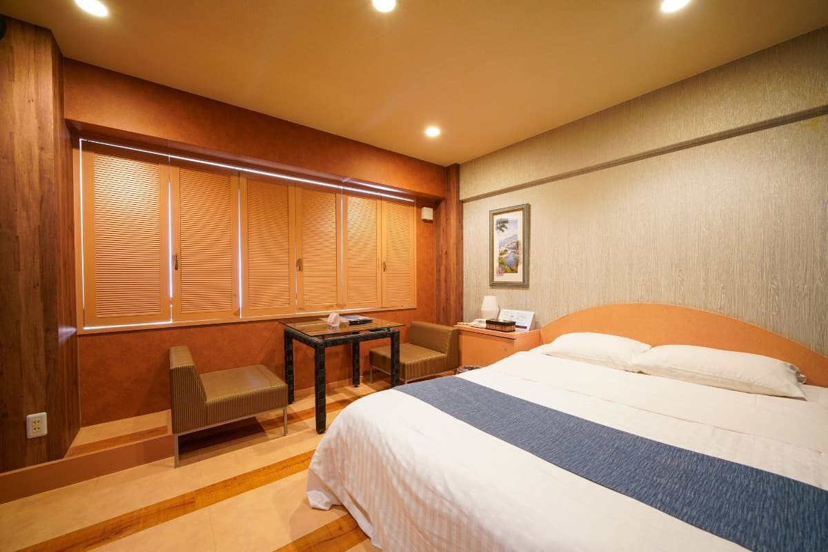 Quarto de hotel com cama, escrivaninha, cadeira e janela com persianas.