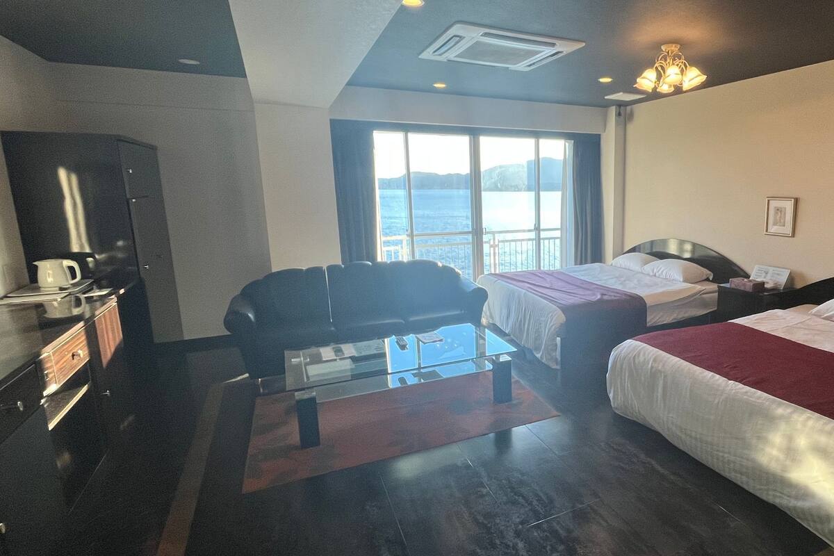 Quarto de hotel com duas camas, um sofá, uma mesa de centro de vidro, uma escrivaninha com abajur e vista para o mar.