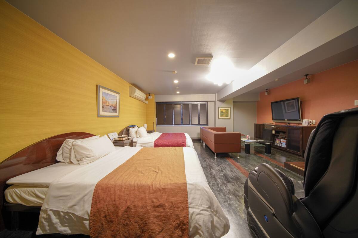 Quarto de hotel com duas camas, um sofá, uma televisão e uma escrivaninha com computador.