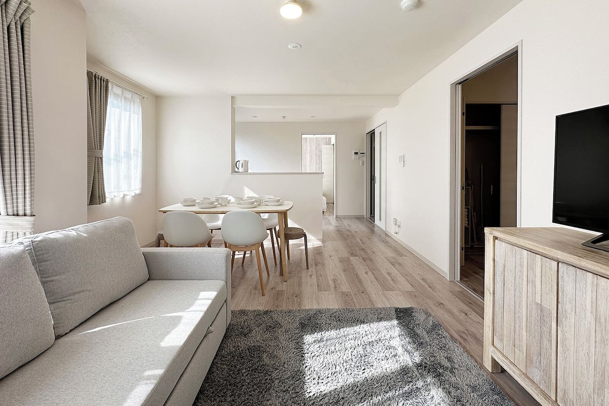 Ein modernes, helles Wohnzimmer mit einer Couch, einem Esstisch mit Stühlen, einem Fernseher auf einem Holzschrank und einem Flur, der zum Schlafzimmer führt.