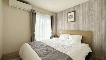 Habitación de hotel con cabecera de madera, cama con ropa de cama blanca y gris, ventana con cortinas translúcidas y un aire acondicionado de pared.