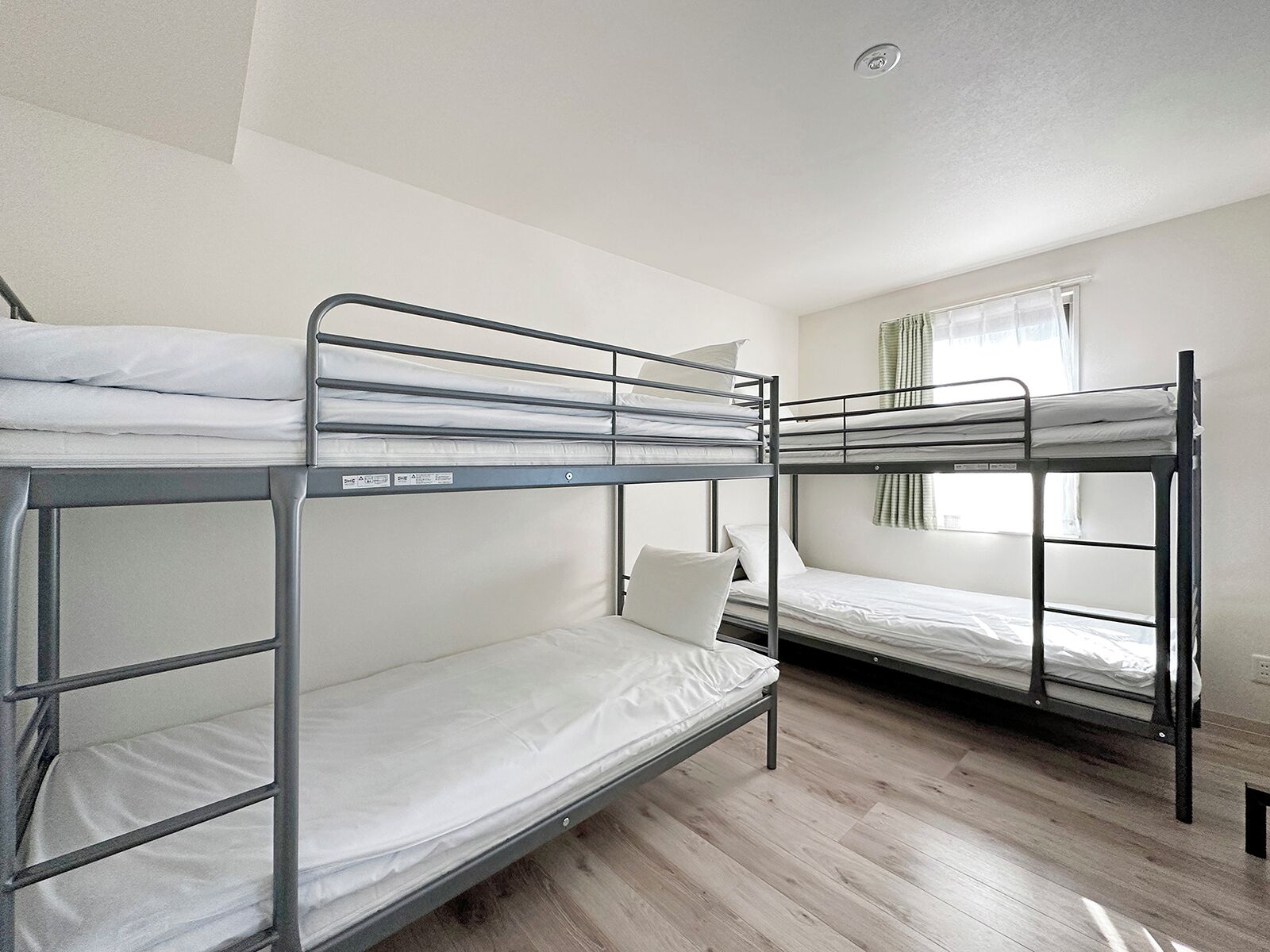 A dormitory room with bunk beds, wooden flooring, and a window with curtains.