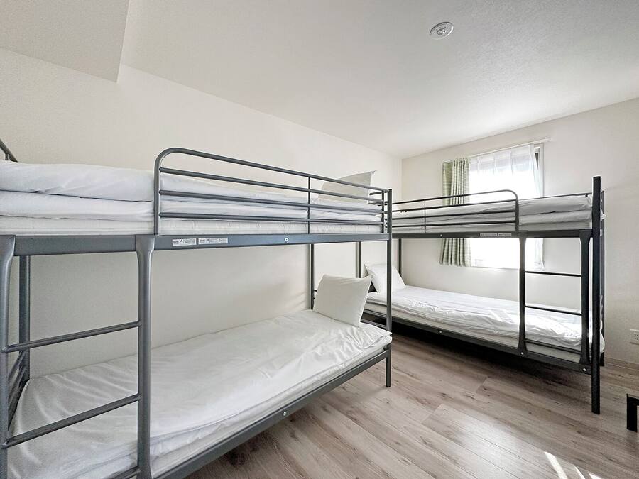 A dormitory room with bunk beds, wooden flooring, and a window with curtains.