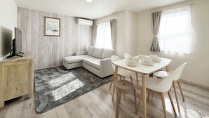A modern living room with a sectional sofa, a dining table with chairs, a TV on a wooden cabinet, and a large window with curtains. - Yutori (Onomichi)