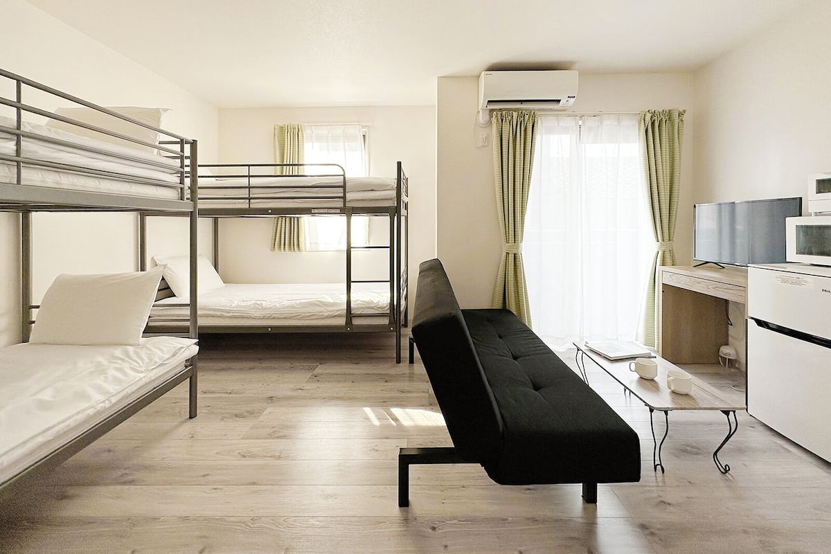 Ein Schlafsaalzimmer mit Etagenbetten, einer schwarzen Schlafcouch, einem Schreibtisch mit Computer und einer Klimaanlage.