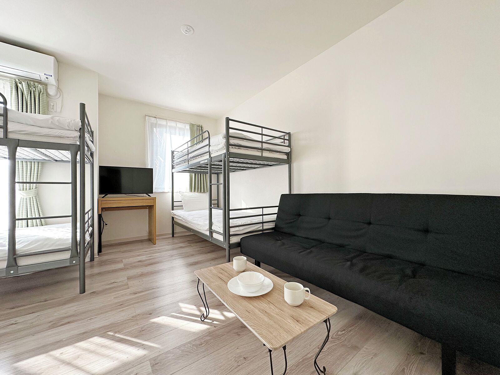 Una habitación tipo dormitorio con literas, un escritorio, un sofá negro y una pequeña mesa con una taza y un platillo.