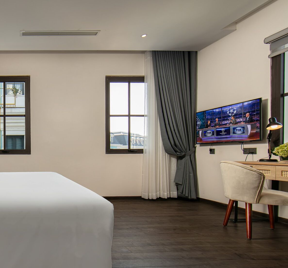 A hotel room with a bed, a desk with a chair, a TV on the wall, and a window with curtains.