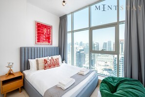 Une chambre moderne avec une grande fenêtre, un lit recouvert de draps blancs, une tête de lit grise et un tableau abstrait rouge.