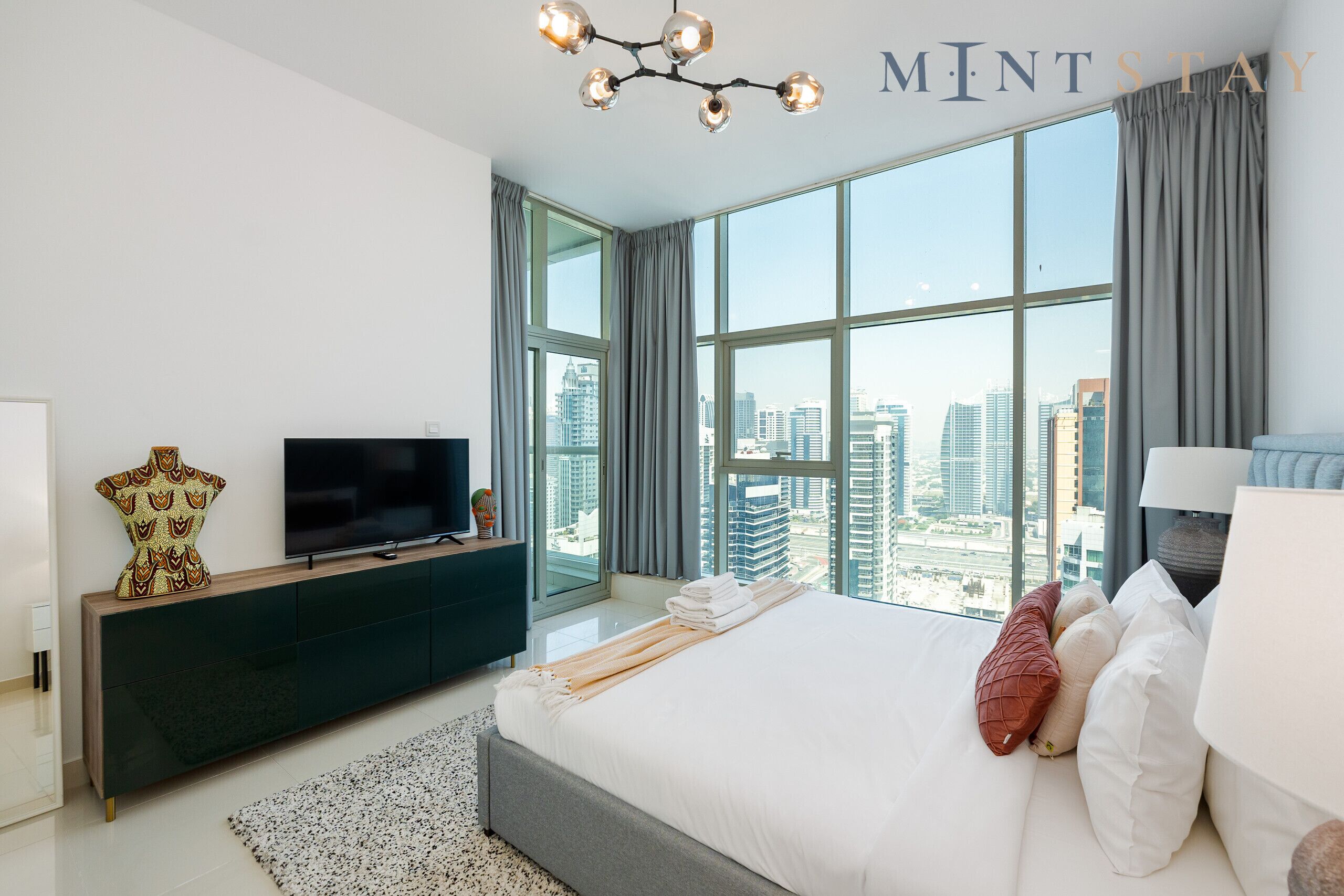 A modern bedroom with a large bed, a flat-screen TV, a decorative vase, and a city view through large windows.