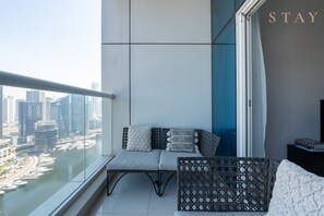 A balcony with a view of a cityscape, a sofa with cushions, a wicker chair, and a small table with a book. - Premium 2BR Apt w/ Marina Views (Dubai)