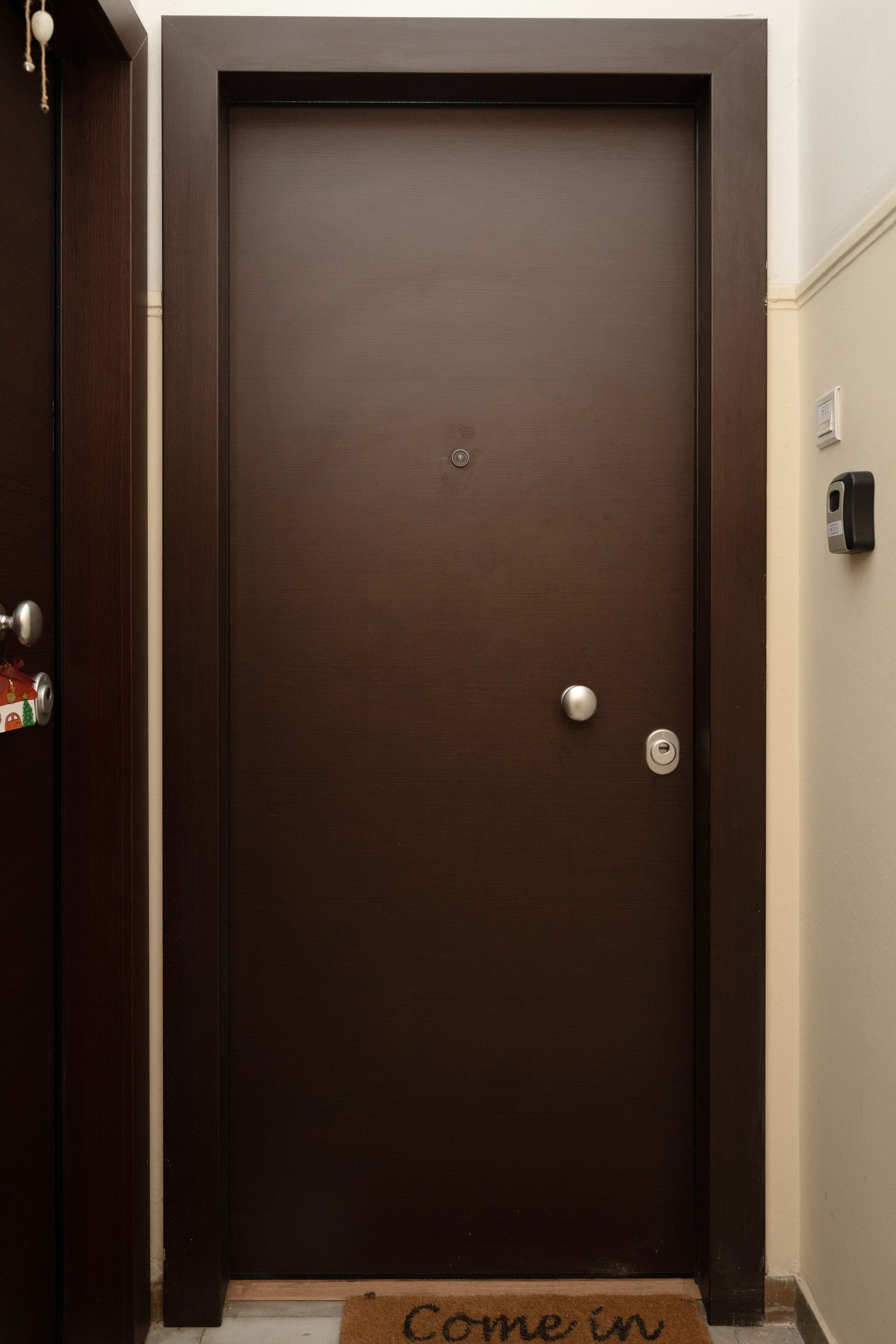 A dark wooden door with a silver handle and a keyhole, a doormat with "Come in" written on it, and a small electronic device mounted on the wall to the right.